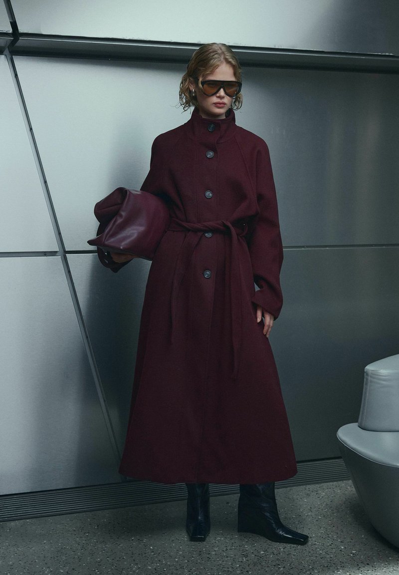 Femme portant un long manteau bordeaux foncé et des bottes noires, tenant un grand sac assorti, debout contre un mur métallique à l'intérieur.