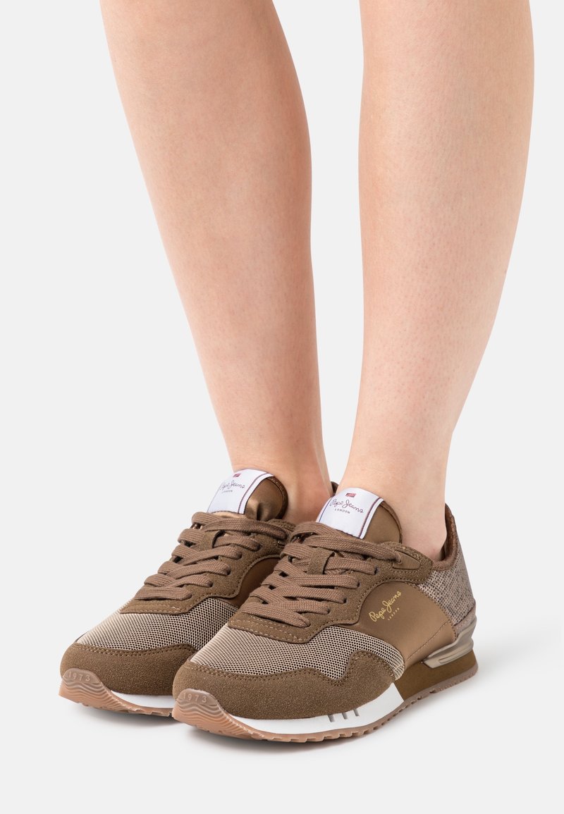 Personne portant des baskets marron à lacets avec des détails en mesh et en daim, debout sur un fond clair.