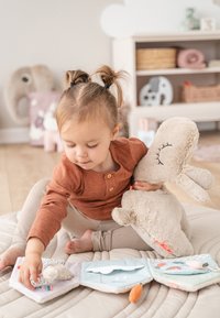 Peuter met vlechtjes speelt met een zacht stoffen boek terwijl ze een grote beige pluchen konijn vasthoudt in een gezellige speelkamer.