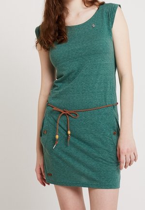 Kobieta w zielonej sukience bez rękawów z brązowym plecionym paskiem związanym w kokardę w talii oraz zapiętymi kieszeniami na guziki.