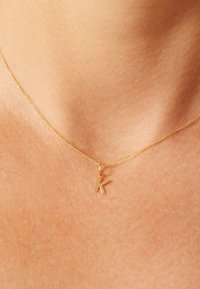 Goldkette mit kleinem Anhänger in Form eines großen Buchstabens "K", vor einem Hautton-Hintergrund präsentiert, der die glatte Oberfläche und den Glanz hervorhebt.