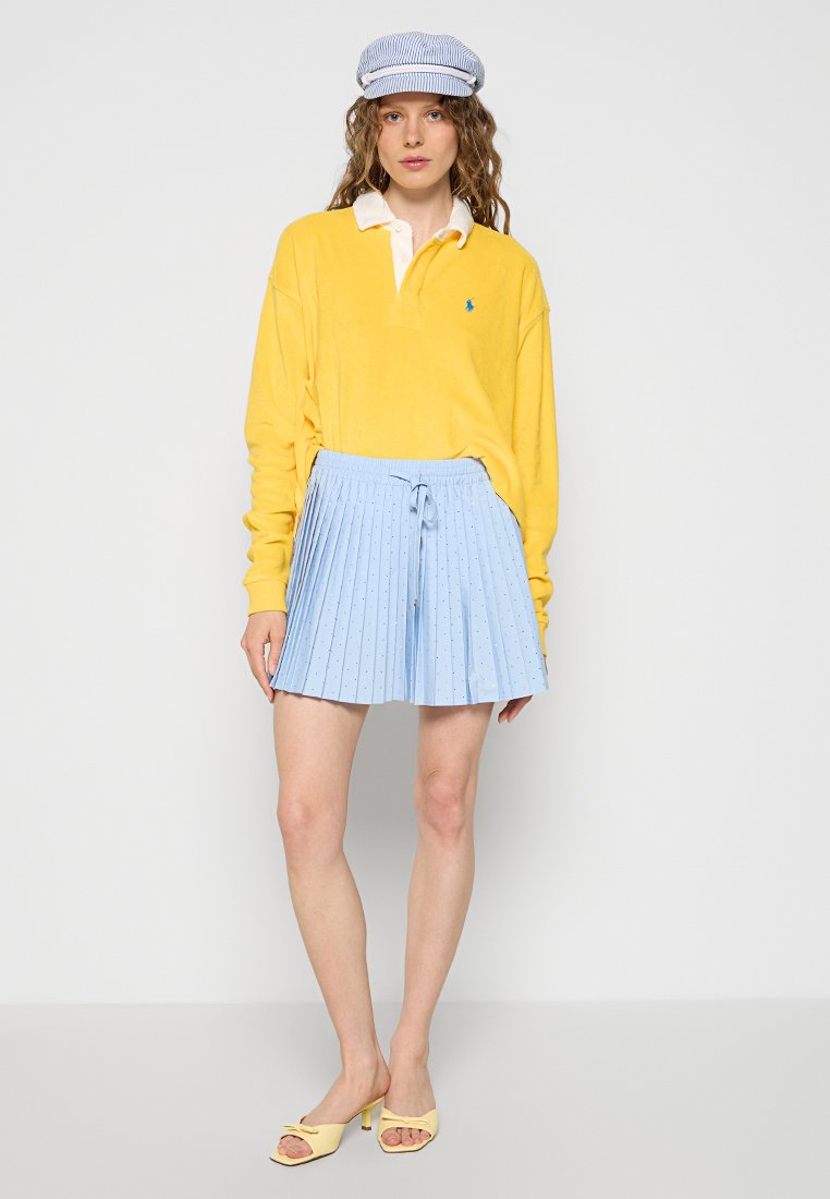 Polo shirt giallo a maniche lunghe, gonna plissettata azzurra chiara con cintura a fiocco, e sandali gialli open-toe. Un cappellino con motivo completa il look.