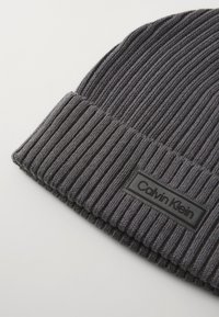 Grå ribbestrikket lue med brettet mansjett og svart rektangulært logo-patch med "Calvin Klein". Stoffet er mykt og elastisk.
