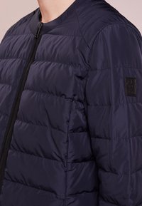 Navy blue puffer jacket with a textured surface, featuring a zip closure and ribbed collar. Includes a small branded patch on the sleeve.
