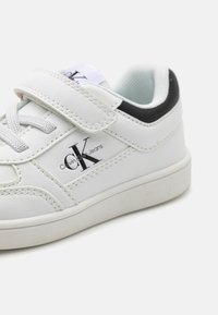 Baskets blanches en cuir avec une sangle Velcro, lacets gris et un accent de talon noir. Présente le logo Calvin Klein Jeans sur le côté. Semelle texturée.