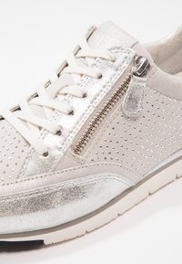 Baskets blanches et argentées avec des côtés perforés, un devant lacé et un détail de fermeture éclair sur le côté sur une semelle blanche.