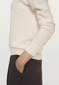 Cremfarbener, gestrickter Pullover mit einem gerippten Saum und Bündchen, kombiniert mit dunkelbraunen Hosen. Hand liegt in der Tasche.