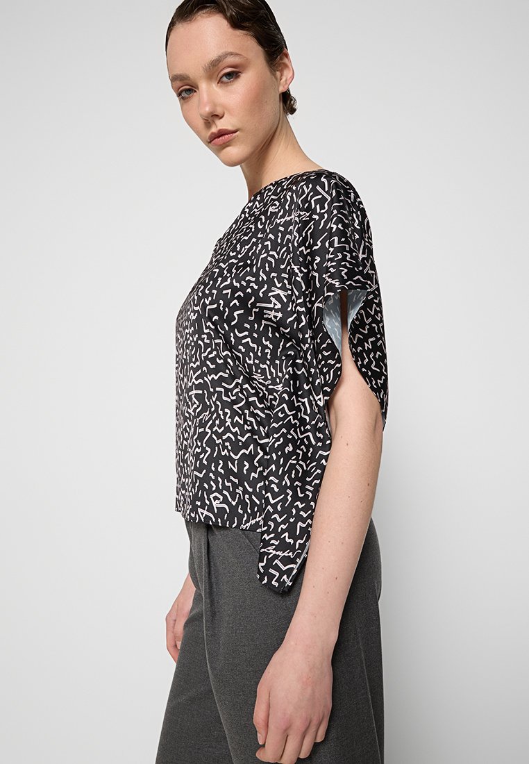 Haut noir avec des motifs abstraits blancs, manches courtes, coupe légèrement ample et couture visible sur l'ourlet. Porté avec un pantalon gris.