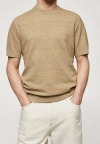 Pull en maille beige à manches courtes avec un col rond, tissu légèrement texturé, associé à un pantalon clair, les mains dans les poches.