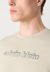 béžové bavlněné tričko s texturovaným šedým logem "calvin klein" na hrudi, kulatým výstřihem a krátkými rukávy.