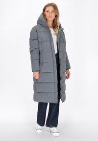 Geacă puffer gri cu glugă, cu fermoar în față, design quilted orizontal și buzunare frontale. Asortată cu blugi închiși și adidași albi.