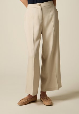 Pantaloni beige a gamba larga con piega frontale, vita alta e bottone decorativo, indossati con mocassini marrone chiaro.