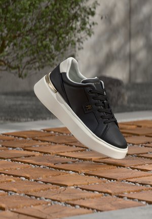 Zapatilla deportiva de cuero blanco y negro con suela blanca gruesa flotando sobre un suelo exterior con baldosas de madera y guijarros.