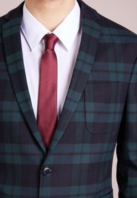 Ruitjes blazer in donkergroen en marineblauw met een satijnen bordeauxrode stropdas, voorzien van een enkele knoopsluiting en een borstzak. Textuurstof.