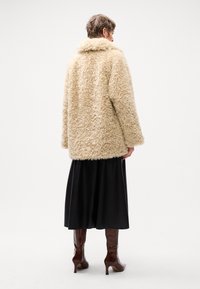 Bež pehme faux fur jakk kõrge kraega, kombineeritud musta satiinse seeliku ja krokodillimustriga pruuni kontsaga saapaga.