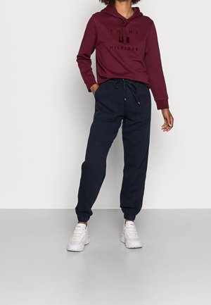 Osoba ubrana w bordową bluzę z kapturem Tommy Hilfiger, granatowe spodnie dresowe ze ściągaczem i białe trampki, stojąca na tle białej, jednolitej ściany.