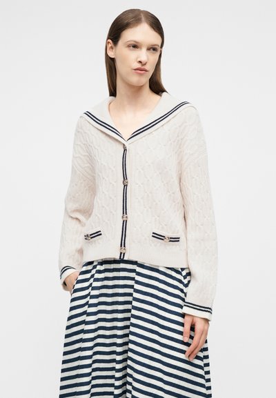 Kvinde iført en hvid kabelstrikket cardigan med marineblå kant og knapper, kombineret med et navy- og hvidstribet nederdel med horisontale striber.