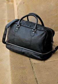 Černá kožená taška přes rameno s kovovými prvky v odstínu stříbra, zipovým uzávěrem, dvojitými držadly na vrchu, odnímatelným ramenním popruhem, hladkou texturou a minimalistickým designem.