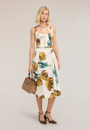 Blondynka w białej sukience midi bez rękawów z żółto-zielonym kwiatowym wzorem, brązowe szpilki, warstwowe naszyjniki i trzymająca brązową torebkę.