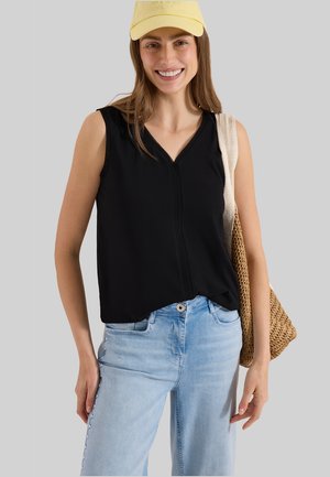 Jeune femme souriante, portant une casquette jaune, un haut noir sans manches, un jean bleu clair, et portant un sac à bandoulière tissé.