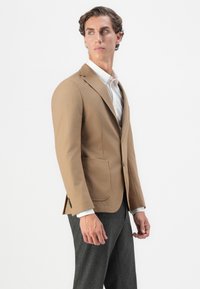 Blazer ajusté marron clair avec des revers à encoches, fermeture à boutons et deux poches avant. Porté sur une chemise blanche, associé à un pantalon gris.