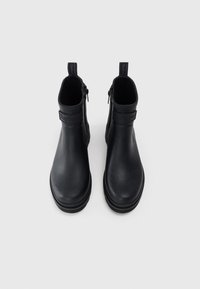 Une paire de bottines noires en cuir avec des zippers sur les côtés et des tirettes marquées "Barbour" sur fond blanc.