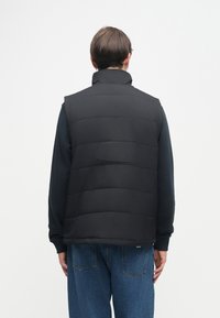 Μαύρο γιλέκο με υψηλό γιακά, που διαθέτει οριζόντια επένδυση. Φορεμένο πάνω από το μακρυμάνικο πουκάμισο και σε συνδυασμό με μπλε τζιν.