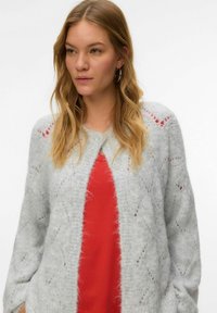 Hellgraue Strickjacke mit weicher Textur, die ein Rautenmuster und rote Akzente aufweist. Knopfverschluss und weite Ärmel. Rotes Oberteil darunter.