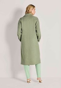 Vrouw in een lange, lichtgroene jas, muntgroene broek en witte hoge hakken, staande tegenover een effen lichte muur.