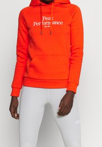 Sweat à capuche orange vif avec poche avant, cordon de serrage à la capuche et logo blanc "Peak Performance". Assorti à des leggings gris clair avec un logo.