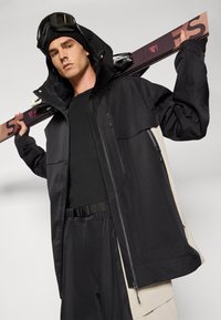 Casaco exterior preto e bege com fecho éclair à frente, grandes bolsos e capuz. O modelo segura skis, vestindo uma camisola e calças de base pretas.