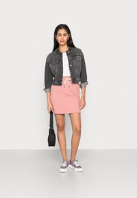 Jaqueta de ganga preta, top branco cropped, saia clara com cordão rosa e ténis estampados. A segurar uma mala preta de ombro.