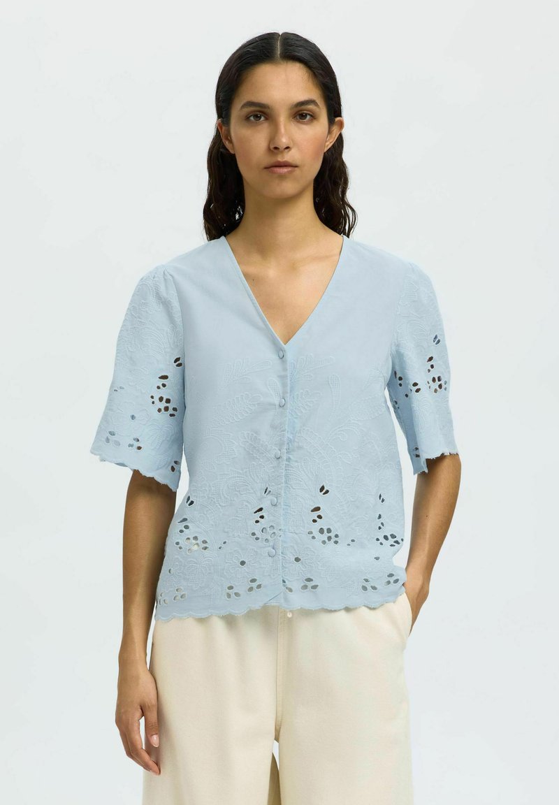 Blusa azzurro chiaro a maniche corte con scollatura a V, caratterizzata da delicati motivi floreali ritagliati, orlo ondulato e bottoni lungo la parte anteriore.