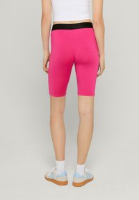 Růžové upnuté cyklistické šortky s černým pásem, hladká textura, délka do poloviny stehna, v kombinaci s bílými ponožkami a teniskami s modrými akcenty.
