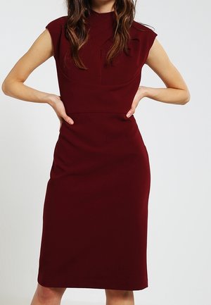 Frau in einem enganliegenden, ärmellosen Midikleid in Burgunderrot mit hohem Ausschnitt, steht mit den Händen in der Hüfte vor einem schlichten Hintergrund.