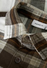 Pruun ja valge ruuduline särk, millel on houndstooth muster, nööbiga kinnitamine ja sildiga, mis ütleb "nimi see." Tekstuuriga kangas ja kaeluse detail.