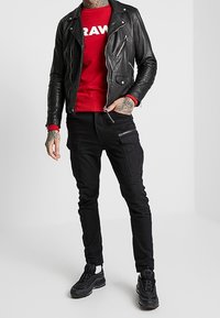 Giacca da biker in pelle nera, felpa rossa con logo "RAW", pantaloni cargo neri e sneakers nere con dettagli bianchi. Vista a figura intera.