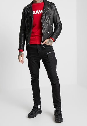 Giacca da biker in pelle nera, felpa rossa con logo "RAW", pantaloni cargo neri e sneakers nere con dettagli bianchi. Vista a figura intera.