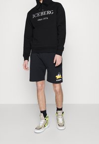 Schwarzer Kapuzenpullover mit dem Text "ICEBERG seit 1974", kombiniert mit schwarzen Shorts mit gelbem Grafikdesign und hochwertigen Sneakers mit Akzenten.