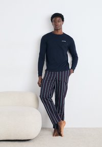 Mørkeblå langærmet skjorte med logo, parret med stripede bukser i rød, grøn og blå. Modellen står barefodet ved siden af en buet, lysefarvet sofa.