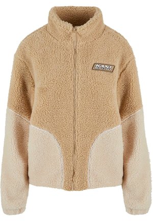 Veste en polaire zippée couleur beige, avec un col montant, un tissu texturé et des panneaux latéraux beige clair contrastants avec un piping blanc. Écusson du logo KANI SPORT.