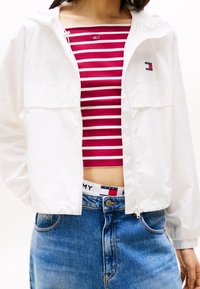 Witte jas met ritssluiting en capuchon, voorzien van twee voorzakken. Daaronder een rood-wit gestreept crop top. Gecombineerd met blauw, high-waisted spijkerbroeken.