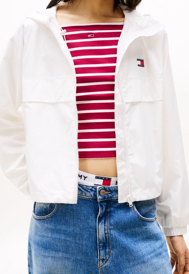 Witte jas met ritssluiting en capuchon, voorzien van twee voorzakken. Daaronder een rood-wit gestreept crop top. Gecombineerd met blauw, high-waisted spijkerbroeken.