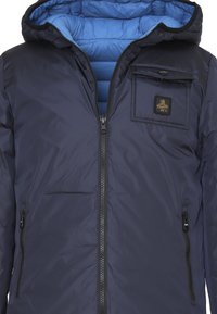 Giubbotto imbottito blu navy con interno blu, chiusura frontale con zip, tasca con zip e patch con logo. Tessuto liscio con cappuccio per maggiore calore.