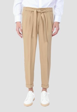 Pantaloni beige a vita alta con taglio affusolato, cintura annodata, orli risvoltati, indossati con camicia bianca e sneaker bianche su sfondo grigio.