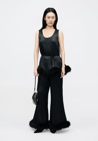 Top negru din satin cu talie reglabilă cu șnur, asortat cu pantaloni evazați cu margine din blană neagră. Modelul ține o geantă mică neagră.