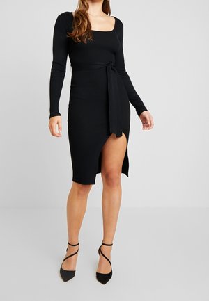 Vrouw die een zwarte, geribbelde jurk met lange mouwen draagt, voorzien van een riem en een hoge split tot op de dij, gecombineerd met zwarte pumps met spitse neus en enkelbandjes.