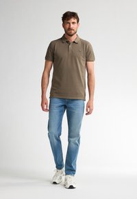 Olivově zelená polokošile s krátkými rukávy, kombinovaná se světle modrými džíny a bílými teniskami. Jednoduchý design bez vzorů nebo akcentů.