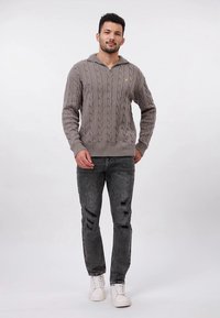 Szary sweter z dzianiny w wzór warkocza z kołnierzem na pół zamek, zestawiony z czarnymi, przetartymi dżinsami i białymi trampkami, model stoi na jednolitym tle.