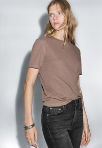 Brown crew-neck t-shirt made of soft fabric, featuring short sleeves, a relaxed fit, and complemented by dark denim jeans. Silvery bangle worn.